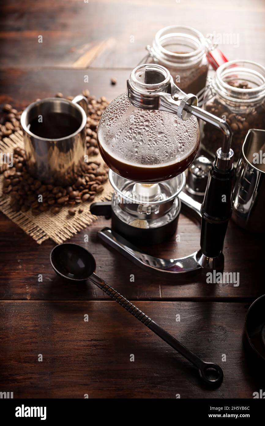 Vacuum coffee maker also known as vac pot, siphon or syphon coffee maker. Metallic cup and toasted coffee beans on rustic wooden table Stock Photo