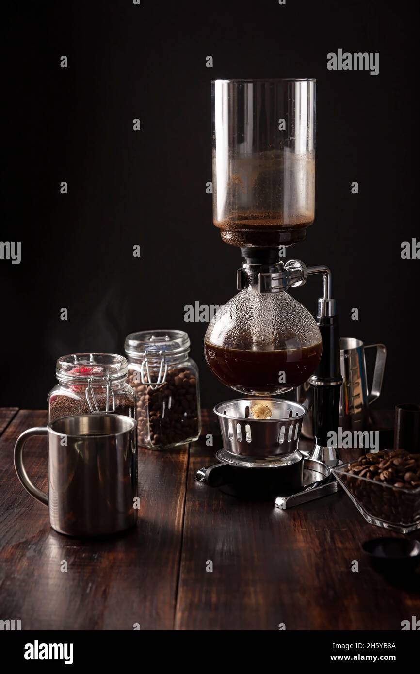 Vacuum coffee maker also known as vac pot, siphon or syphon coffee maker. Metallic cup and toasted coffee beans on rustic wooden table Stock Photo