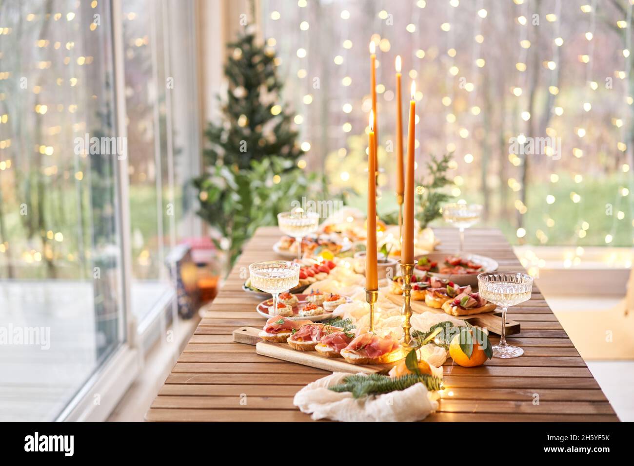 Christmas dinner feast. A small table is served with snacks, bruschettas, and canapes. A decorated dining table with champagne glasses, candles and ch Stock Photo