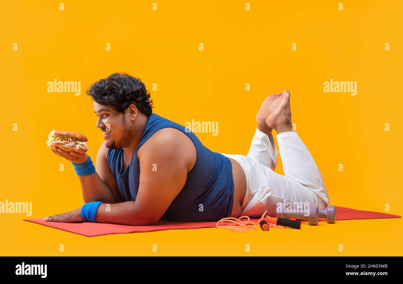 A fat man lying on yoga mat greedily devouring  a burger In hand with dumbles and skipping rope kept beside. Stock Photo