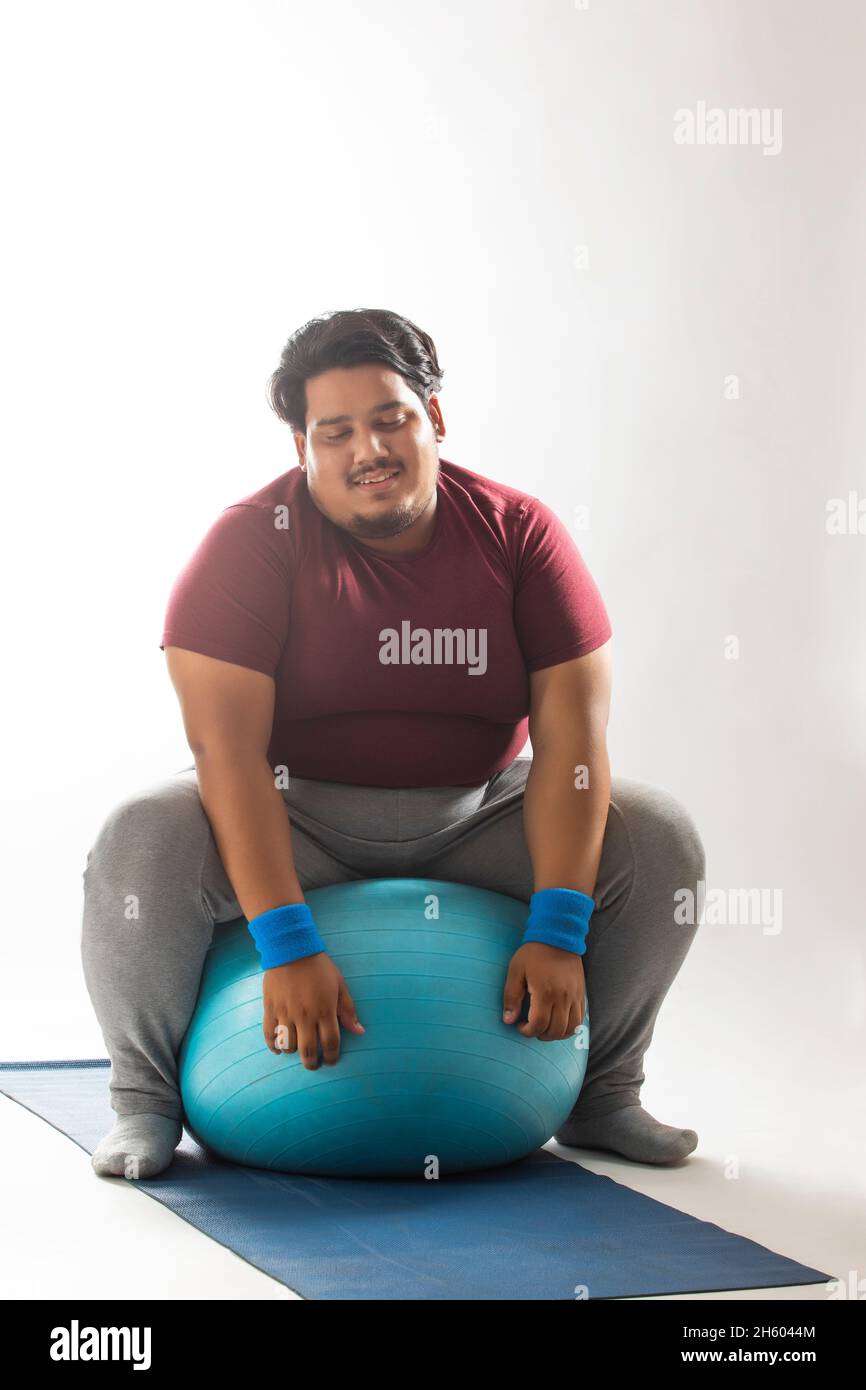 A fat man sitting with closed eyes on a fitness ball kept on yoga mat against plain background. Stock Photo