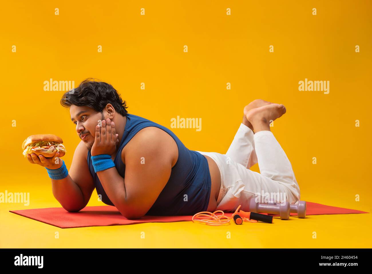 A fat man lying on yoga mat greedily devouring  a burger In hand with dumbles and skipping rope kept beside. Stock Photo