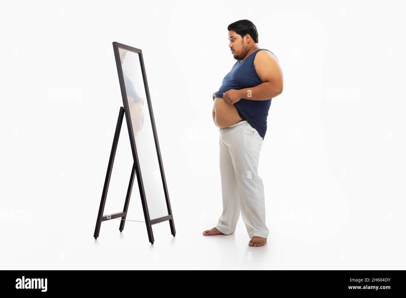 Portrait of a fat man looking at bellyfat in mirror with surprise against plain background. Stock Photo