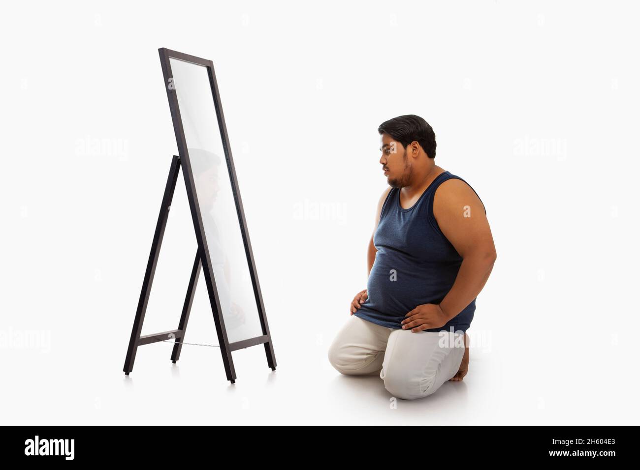 Portrait of a fat man looking at bellyfat mirror in despair against plain background. Stock Photo