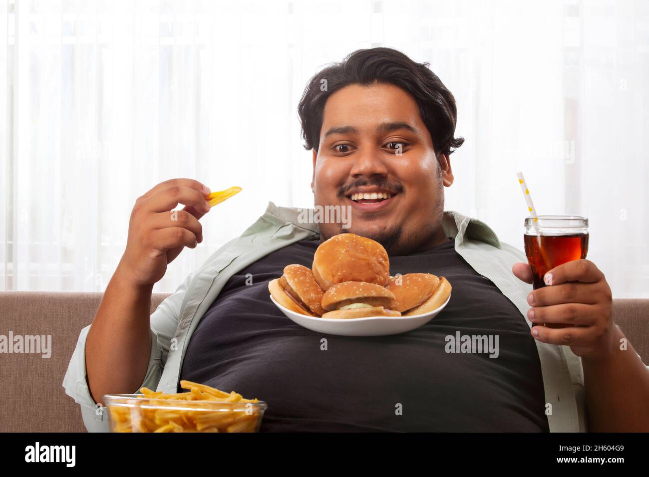 A fat man smiling while sitting in his room with junk food In his hand and on chest. Stock Photo