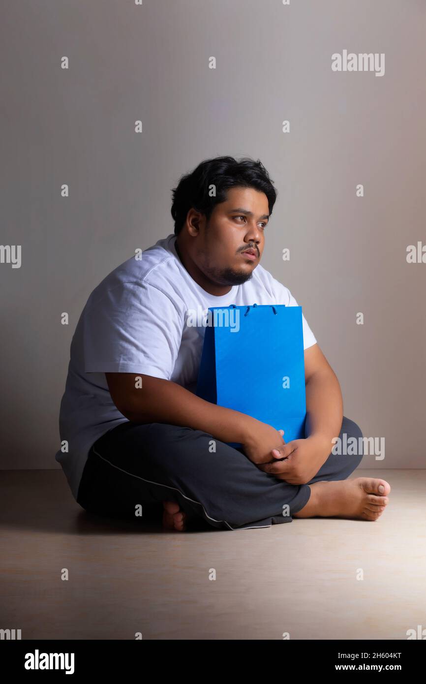 A fat man sitting sadly with a shoppingbag in hand. Stock Photo