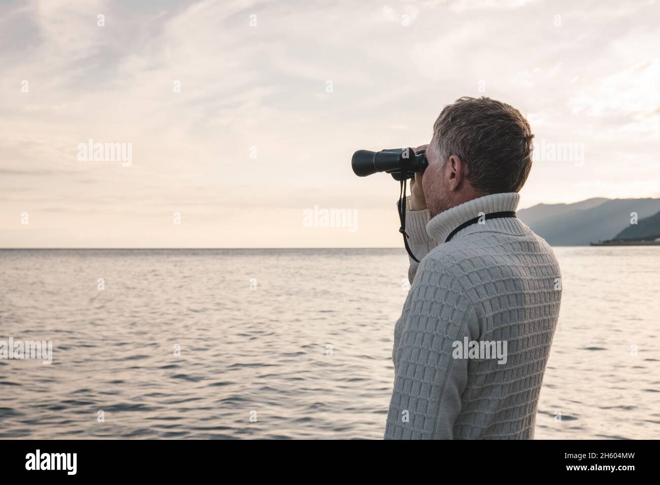 Portrait of a handsome middle-aged man in a white sweater looking through binoculars at the sea in the evening at sunset. Space for text. Stock Photo