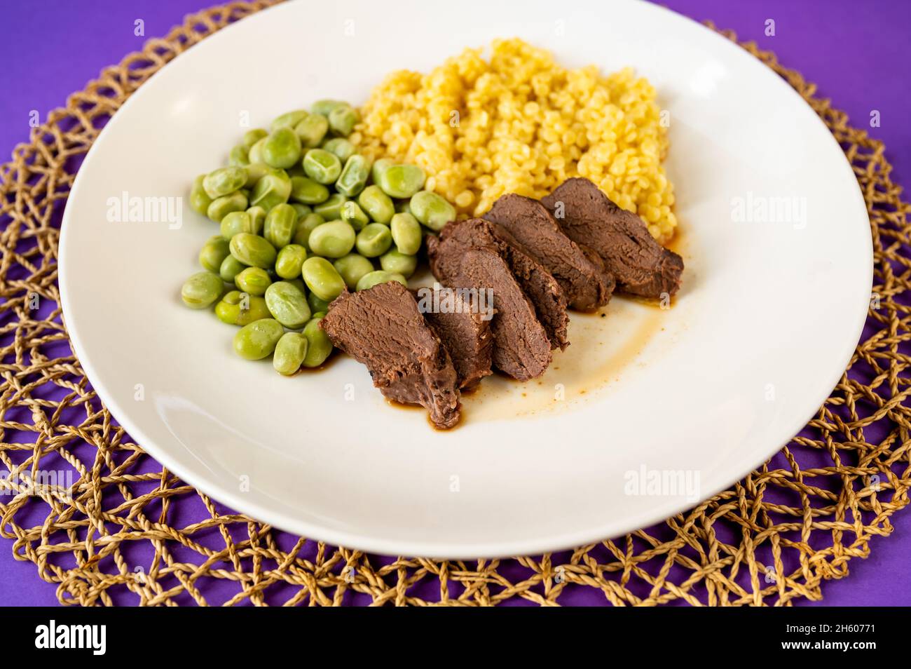 Sliced lean deer meat, boiled broad bean and semolina pasta 'tarhona' on plate on circle bamboo pad on table, closeup. Stock Photo
