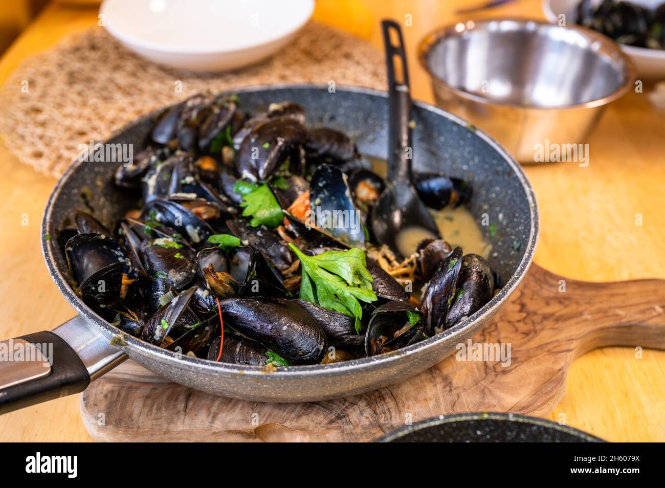 Boiled black mussels in wine and garlic in pan with spoon,bowl and plate on table, closeup. Stock Photo