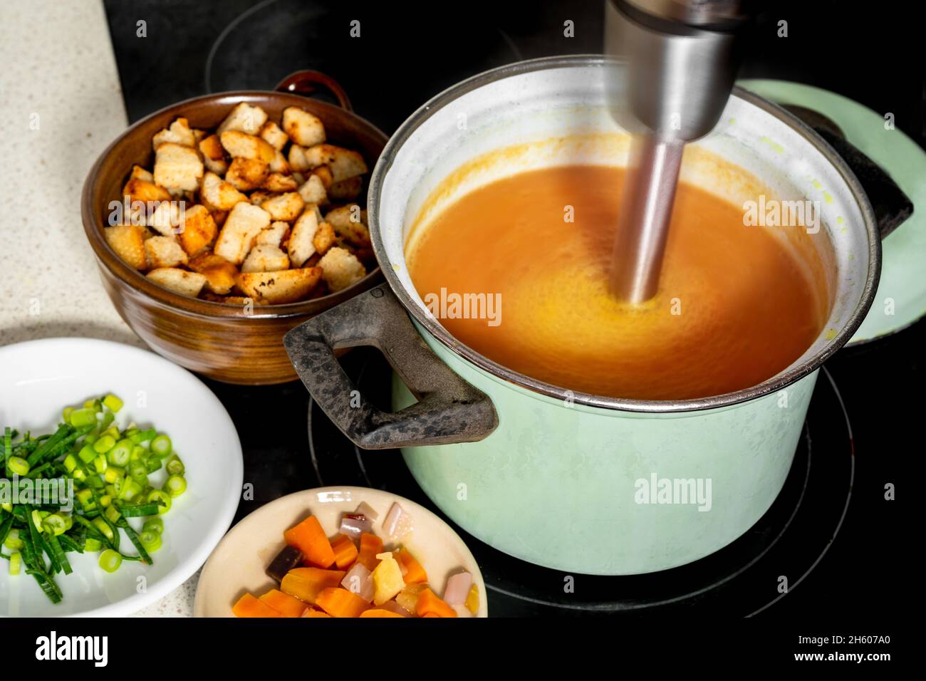 Hand blender in pot on stove mixes carrot soup, plates with vegetable and sliced fried bread on kitchen board. Preparation fresh carrot soup. Stock Photo