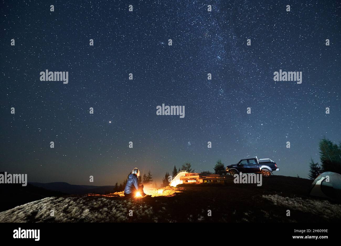 Lonely woman sitting on grass by burning fire near two tents and black SUV. Night camping on mountain hill under starfull sky. Tree tops and silhouettes of mountains afar on the background. Stock Photo