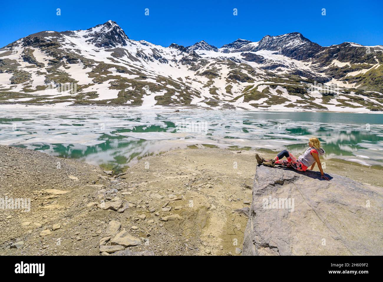 Woman in the snow relaxes sitting by reflecting White lake or Lago Bianco of Switzerland. Lakefront on iced White lake in Grisons Canton at the Stock Photo