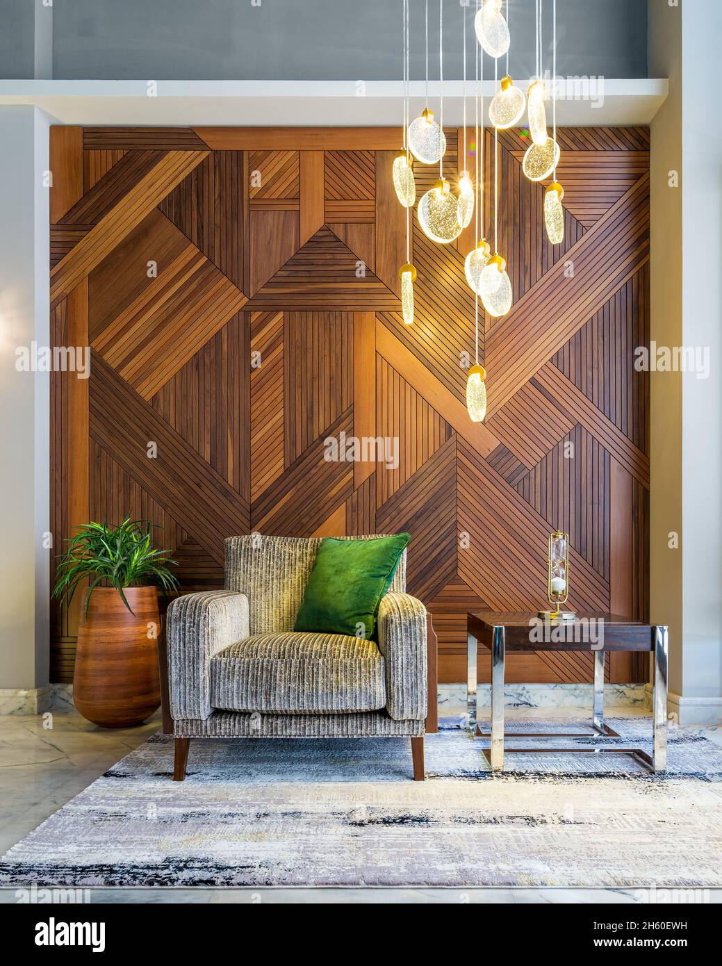 Interior of modern beige armchair, small wooden modern table, planter with green bushes, and contemporary tall glass chandelier, in a hall with decorated wood cladding wall, and white marble floor Stock Photo
