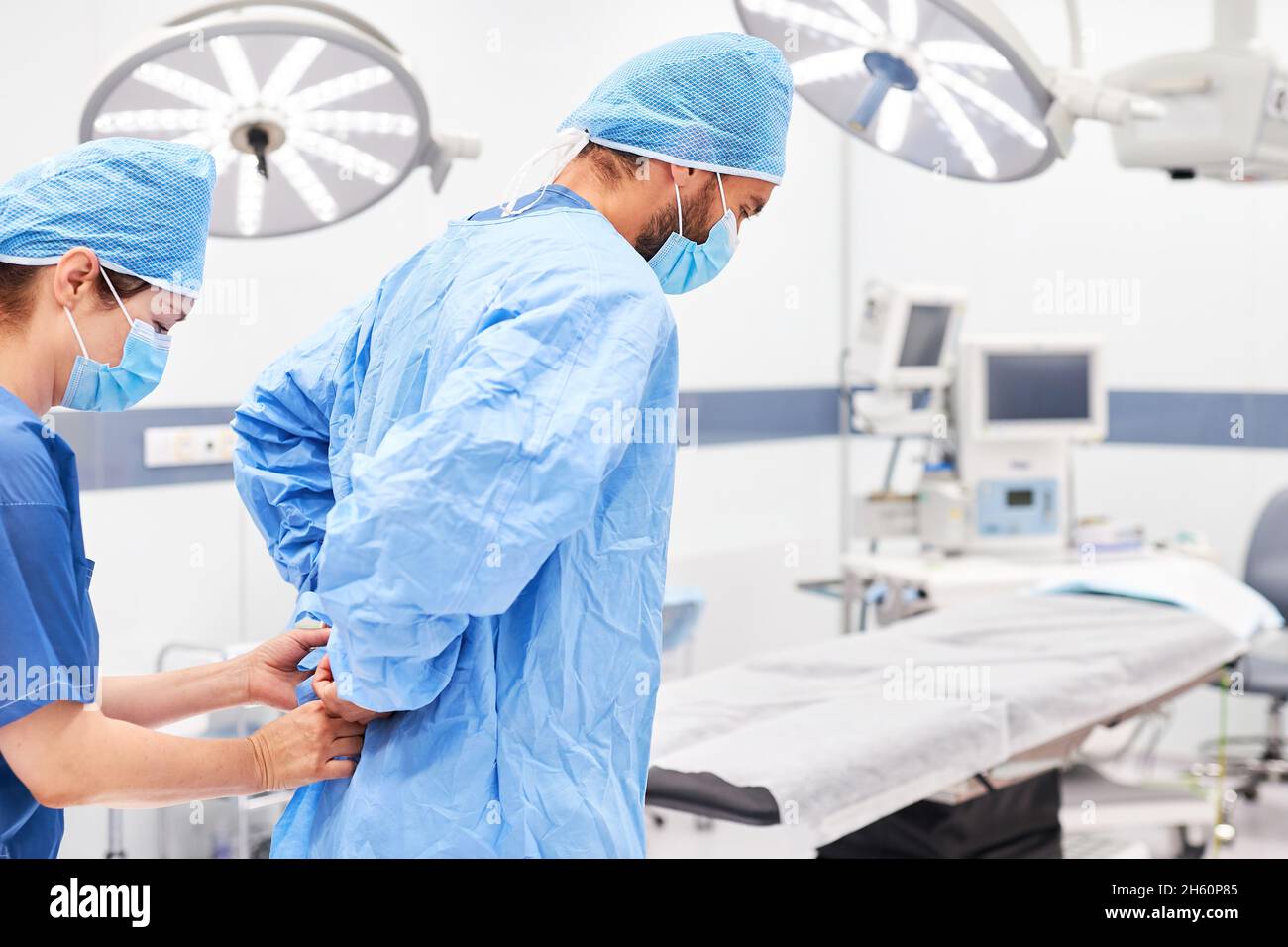 Nurse helps surgeon with face mask put on gown before surgery Stock Photo
