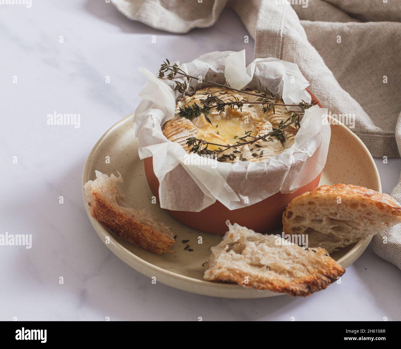 Camembert fondue with thyme in a small terracotta dish placed on a serving plate with pieces of sourdough bread. Stock Photo