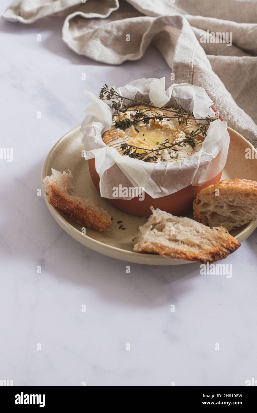 Camembert fondue with thyme in a terracotta dish placed on a serving plate with pieces of sourdough bread. Stock Photo