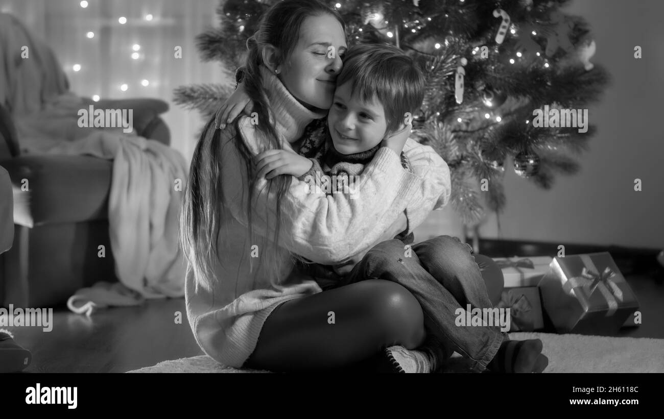 Black and white portrait of happy smiling mother with son hugging next to glowing Christmas tree at home Stock Photo