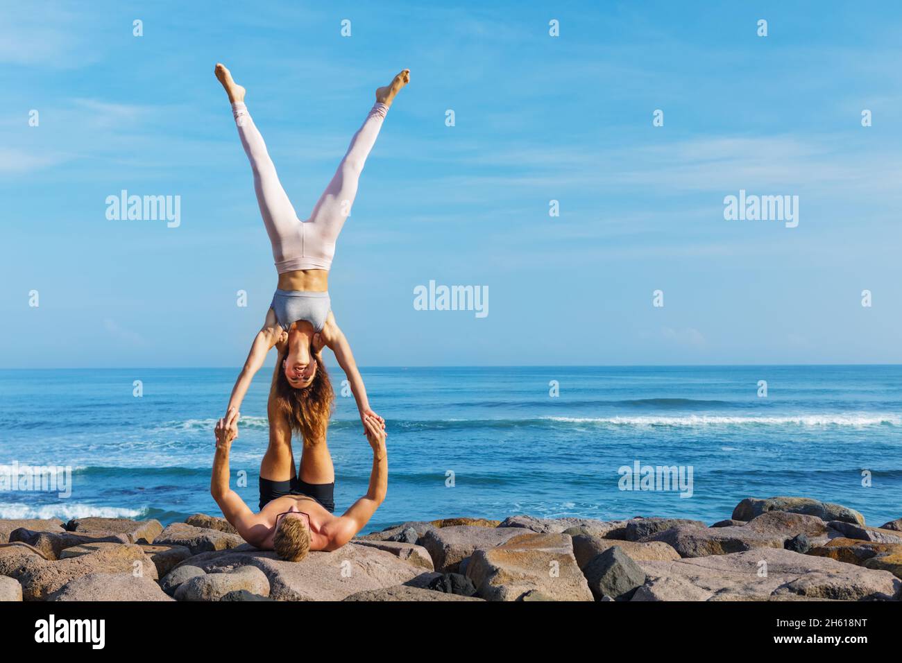 Fit young couple doing acro yoga at spa retreat on sea beach. Active woman on partner feet, balancing at handstand acroyoga pose. Healthy lifestyle. P Stock Photo