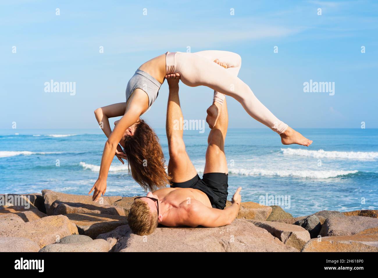 Fit young couple doing acro yoga at spa retreat on sea beach. Active woman balancing on partner feet, stretching at acroyoga pose. Healthy lifestyle. Stock Photo