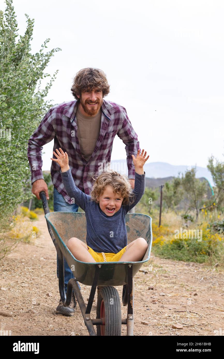 Young man pushing cheerful and excited son sitting with hands raised in wheelbarrow on walkway Stock Photo