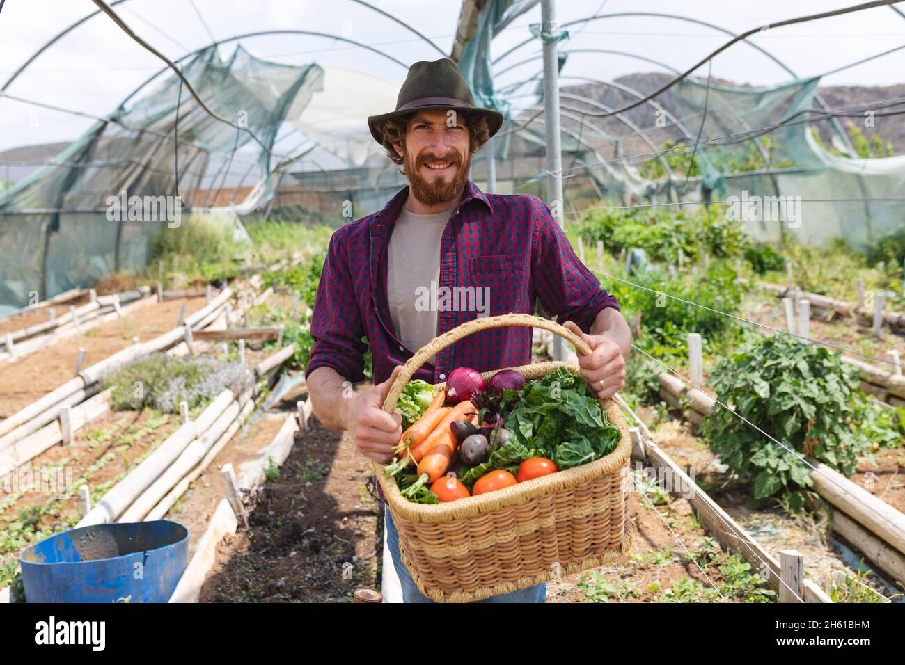 Portrait of smiling bearded man wearing hat carrying freshly harvested vegetables from organic farm Stock Photo