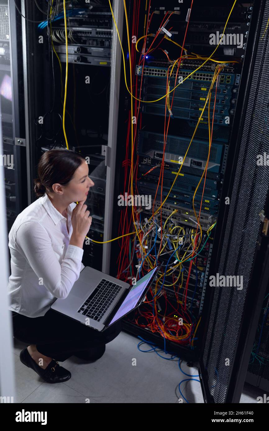 Thoughtful caucasian female engineer with laptop inspecting computer server in computer server room Stock Photo