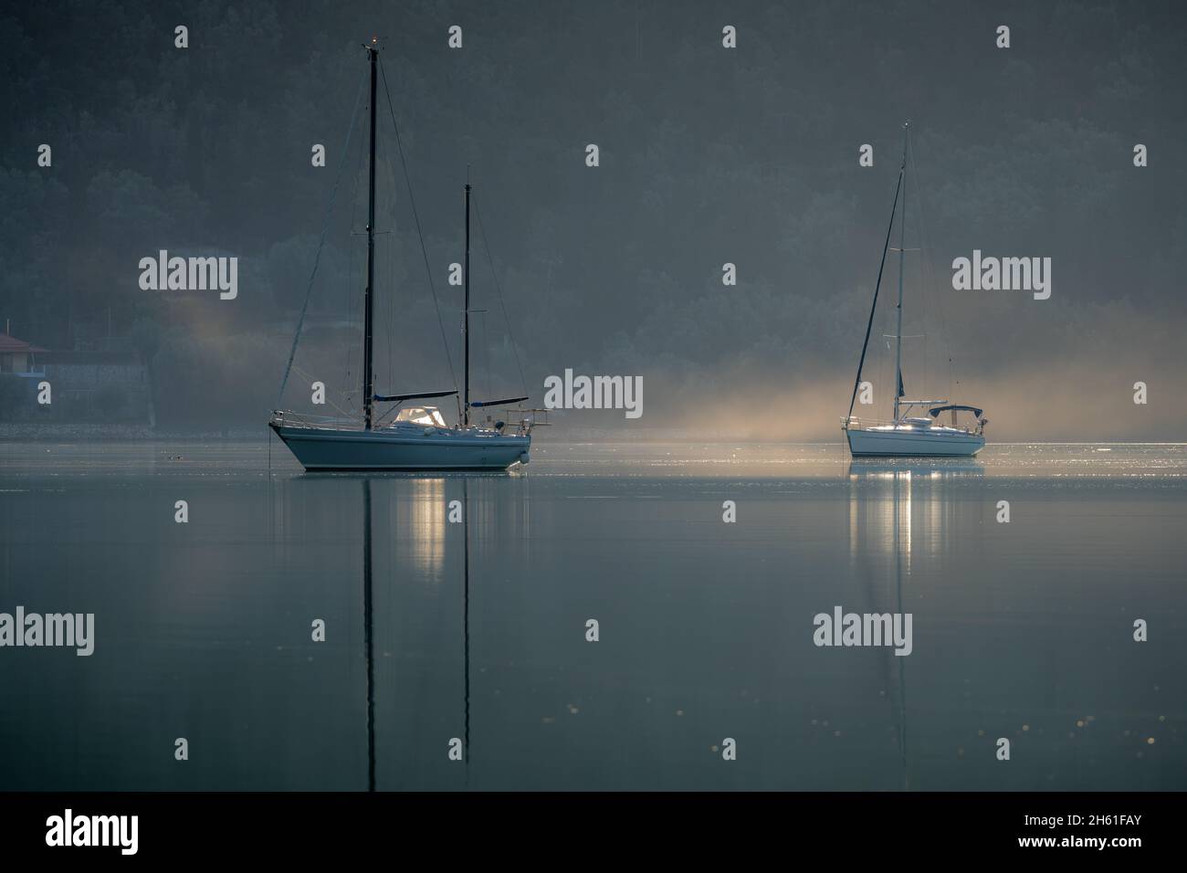 Yachts on the calm water of a sheltered bay casting reflections with morning mist on the water surface. Stock Photo