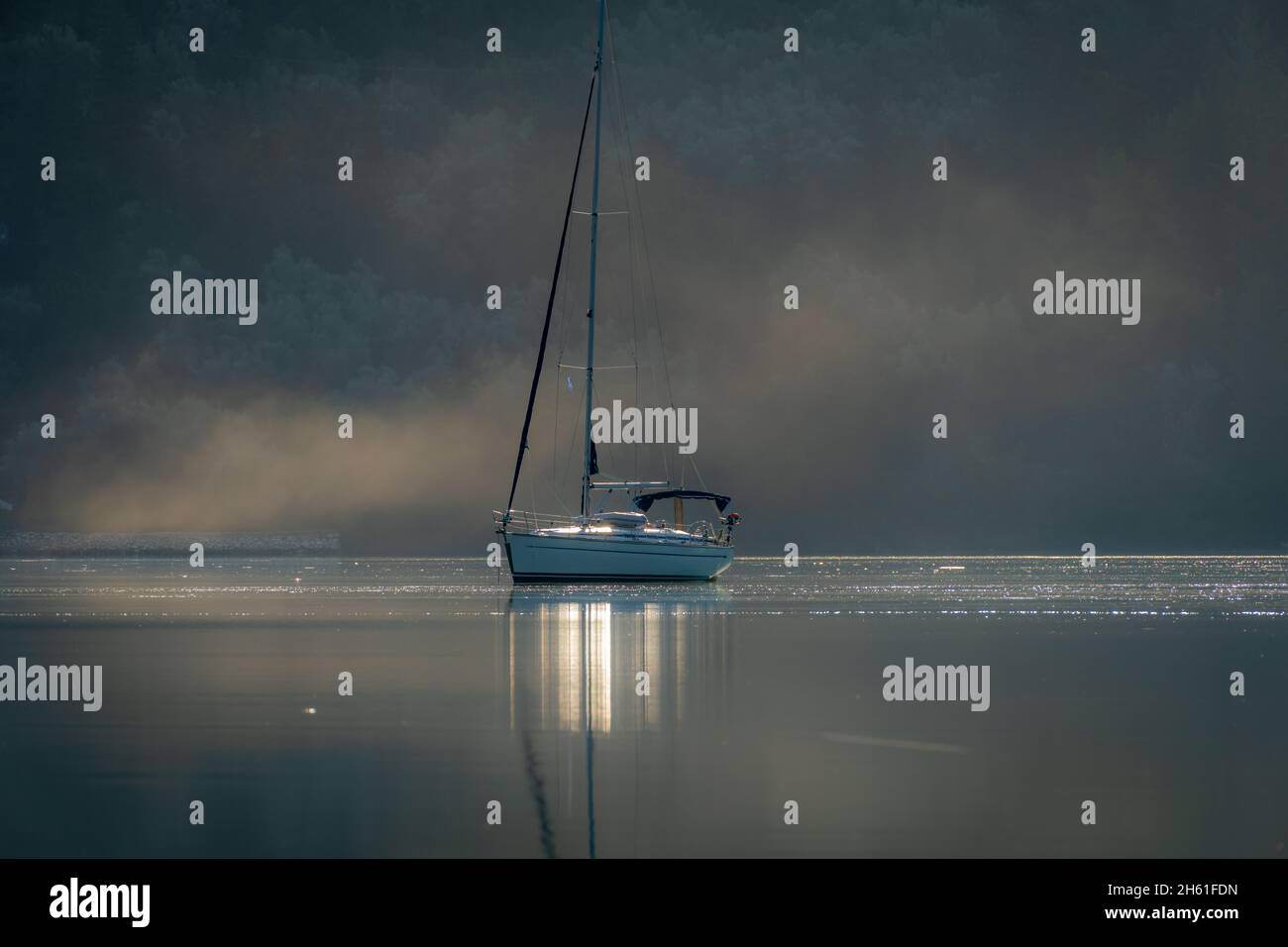 A sailing boat on the calm water of a sheltered bay casting reflections with morning mist on the sea surface. Stock Photo