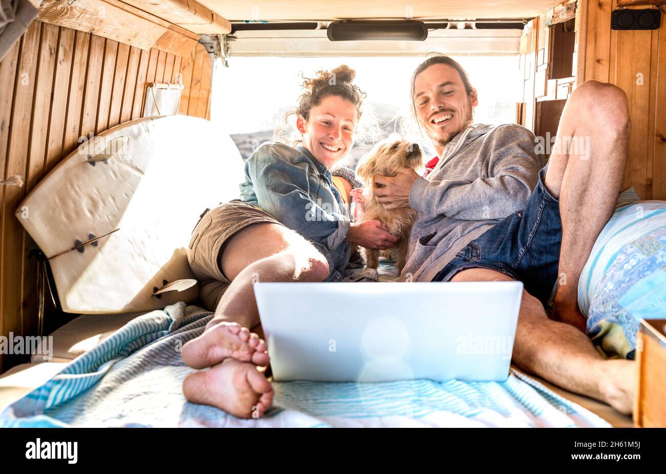 Digital nomad couple with cute dog using laptop on retro mini van transport - Travel life inspiration concept with indie people on minivan Stock Photo