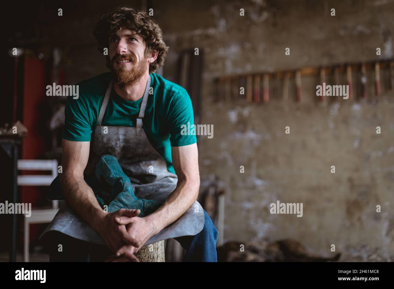 Smiling blacksmith looking away while sitting with hands clasped in metal industry Stock Photo