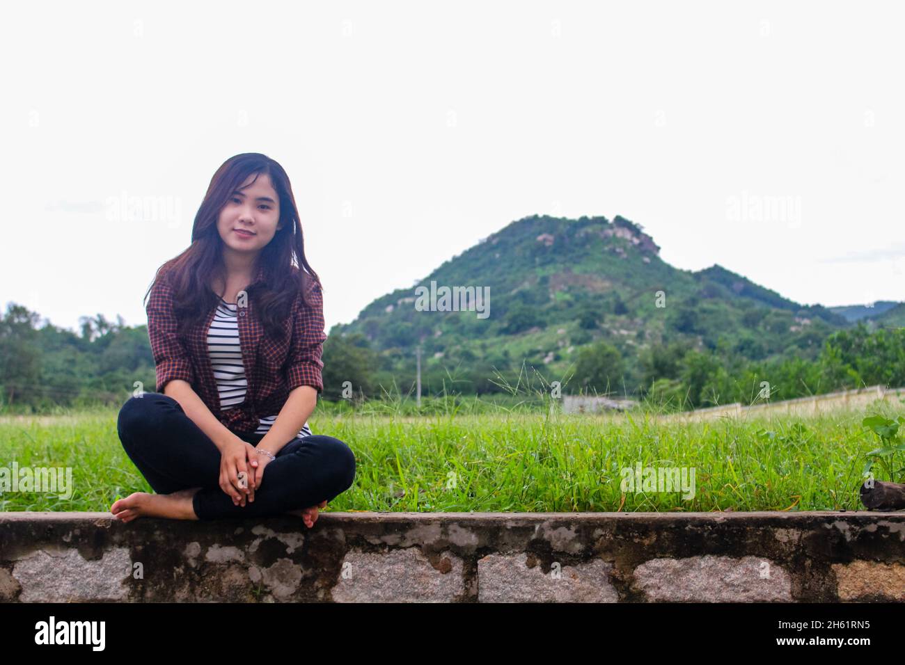 A young beautiful Vietnamese woman is sitting in the grass near a hillside in the sunny day Stock Photo