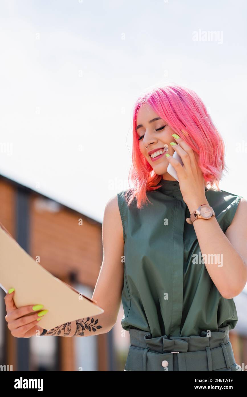 pleased businesswoman with pink hair holding folders and talking on smartphone outside Stock Photo