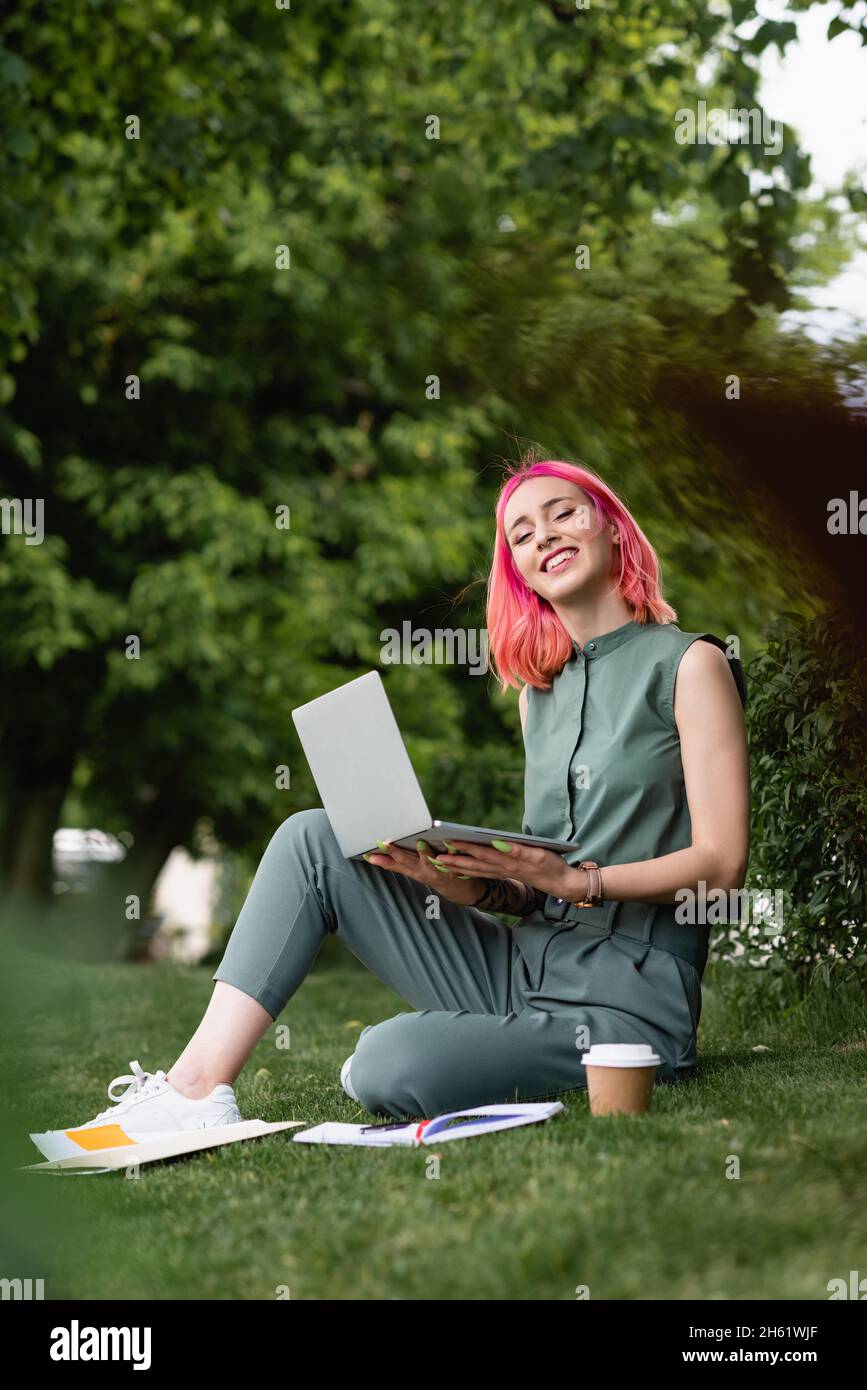 joyful woman with pink hair holding laptop and sitting on grass Stock Photo