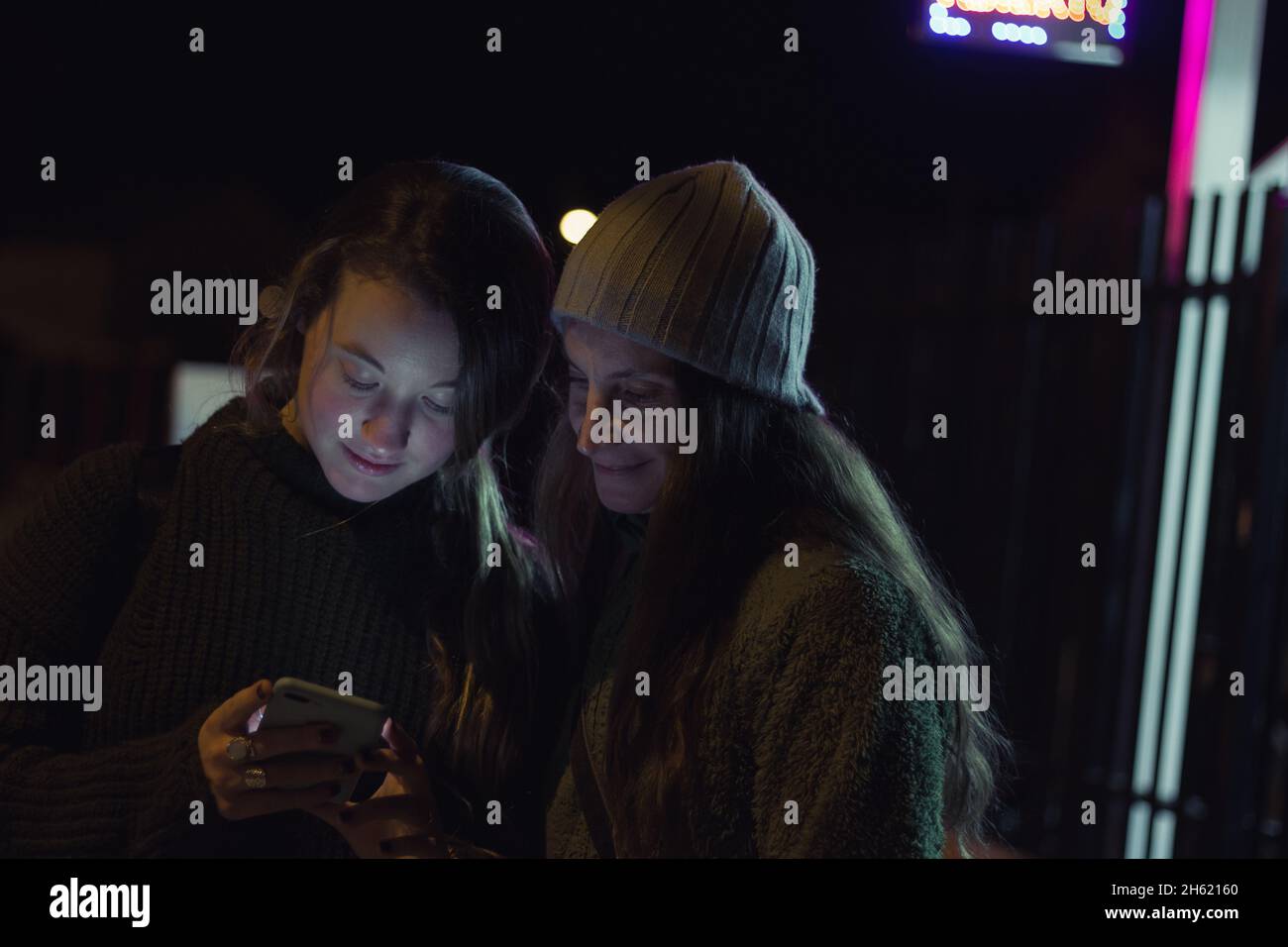 Young girl using cell phone next to older sister with grey knit hat on cold winter night outdoors Stock Photo