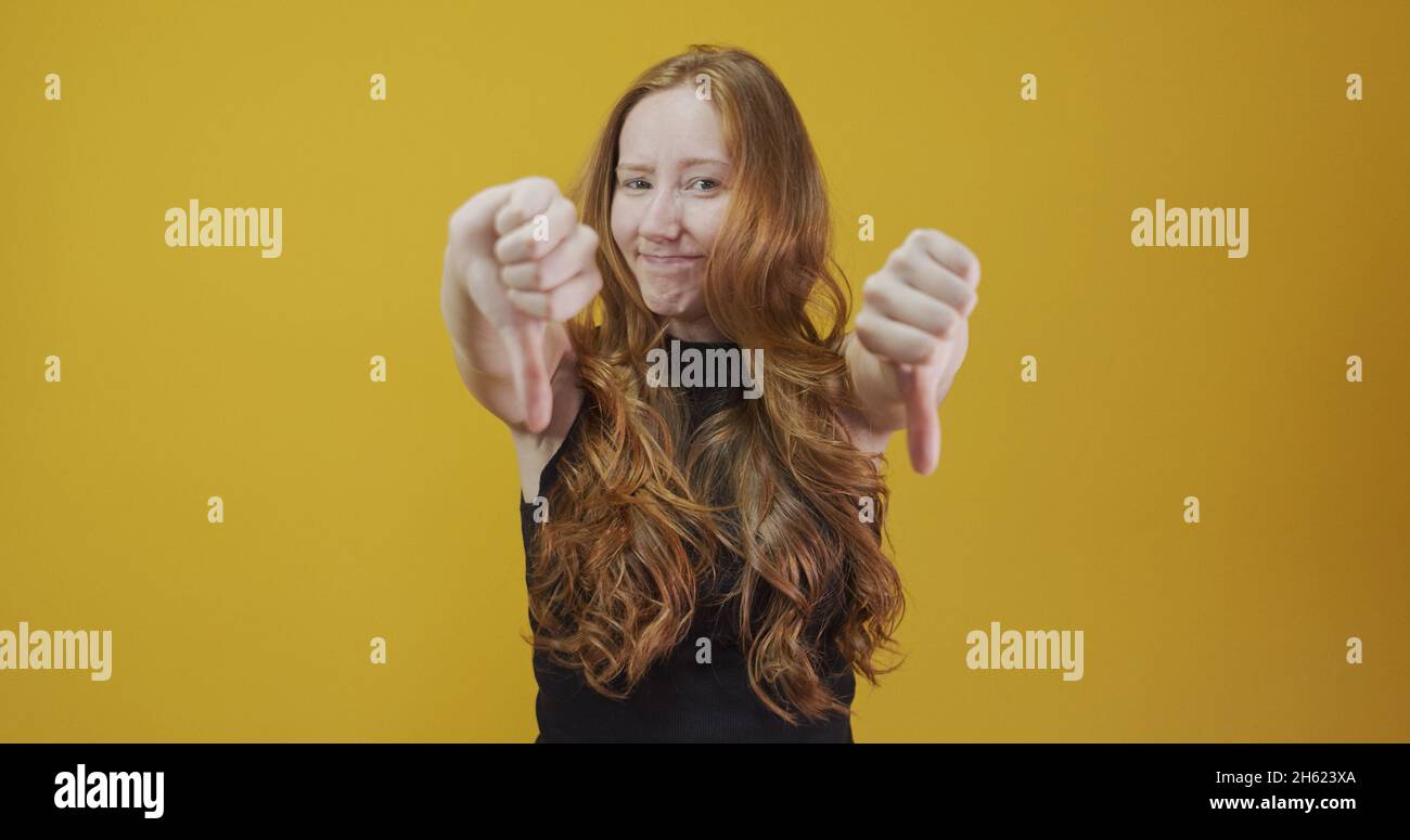 Unhappy redhead woman giving thumbs down gesture looking with negative expression and disapproval. Yellow background. Stock Photo