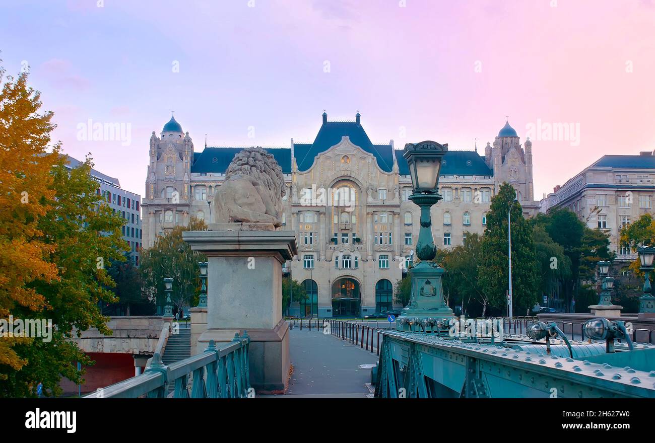The carved stone Gresham Palace facade behind the sculpture of lion and vintage streetlight of the Chain Bridge, against the purple sunrise sky, Budap Stock Photo
