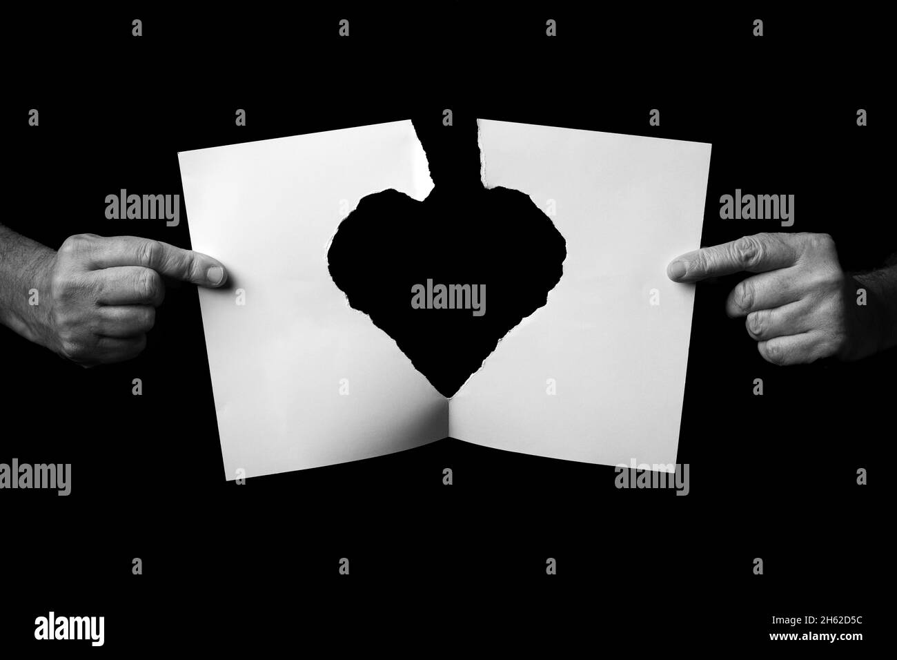 B+W image of male hands holding and ripping sheet of paper with torn heart shape against black background. Stock Photo