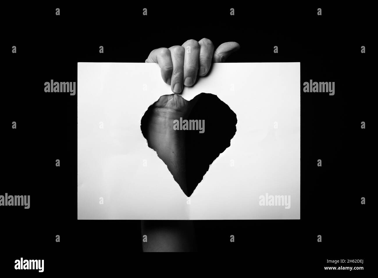 B+W image of male hand holding sheet of paper with torn heart shaped hole against black background. Stock Photo