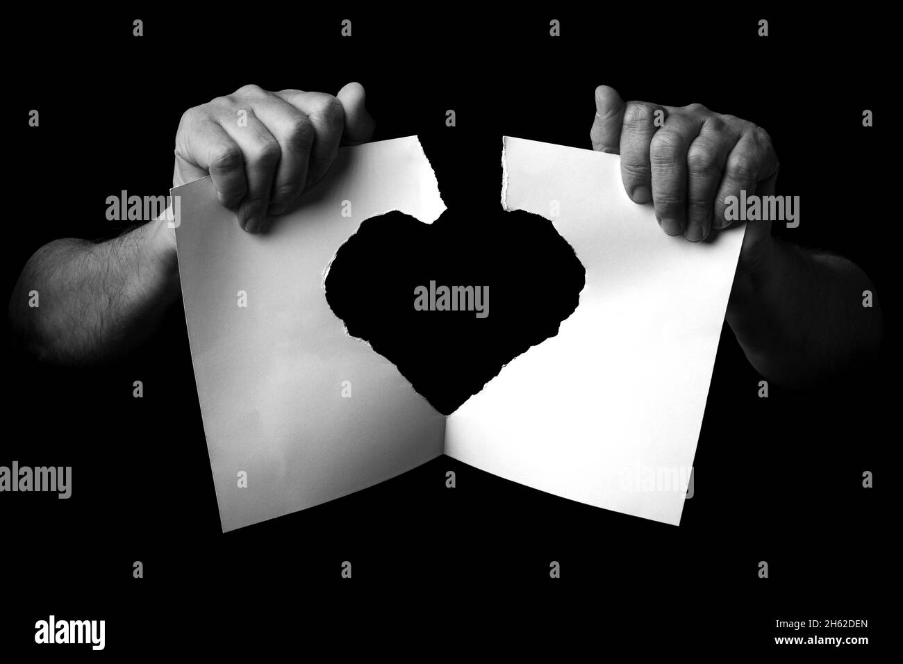 B+W image of male hands holding and ripping sheet of paper with torn heart shape against black background. Stock Photo