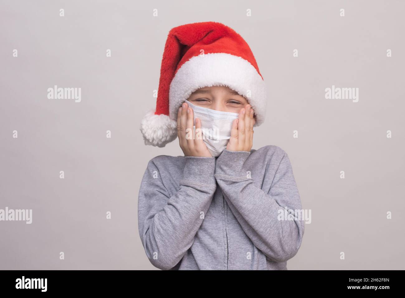 A boy in a Santa helper hat and a medical protective mask holds a mask with his hands.  Stock Photo