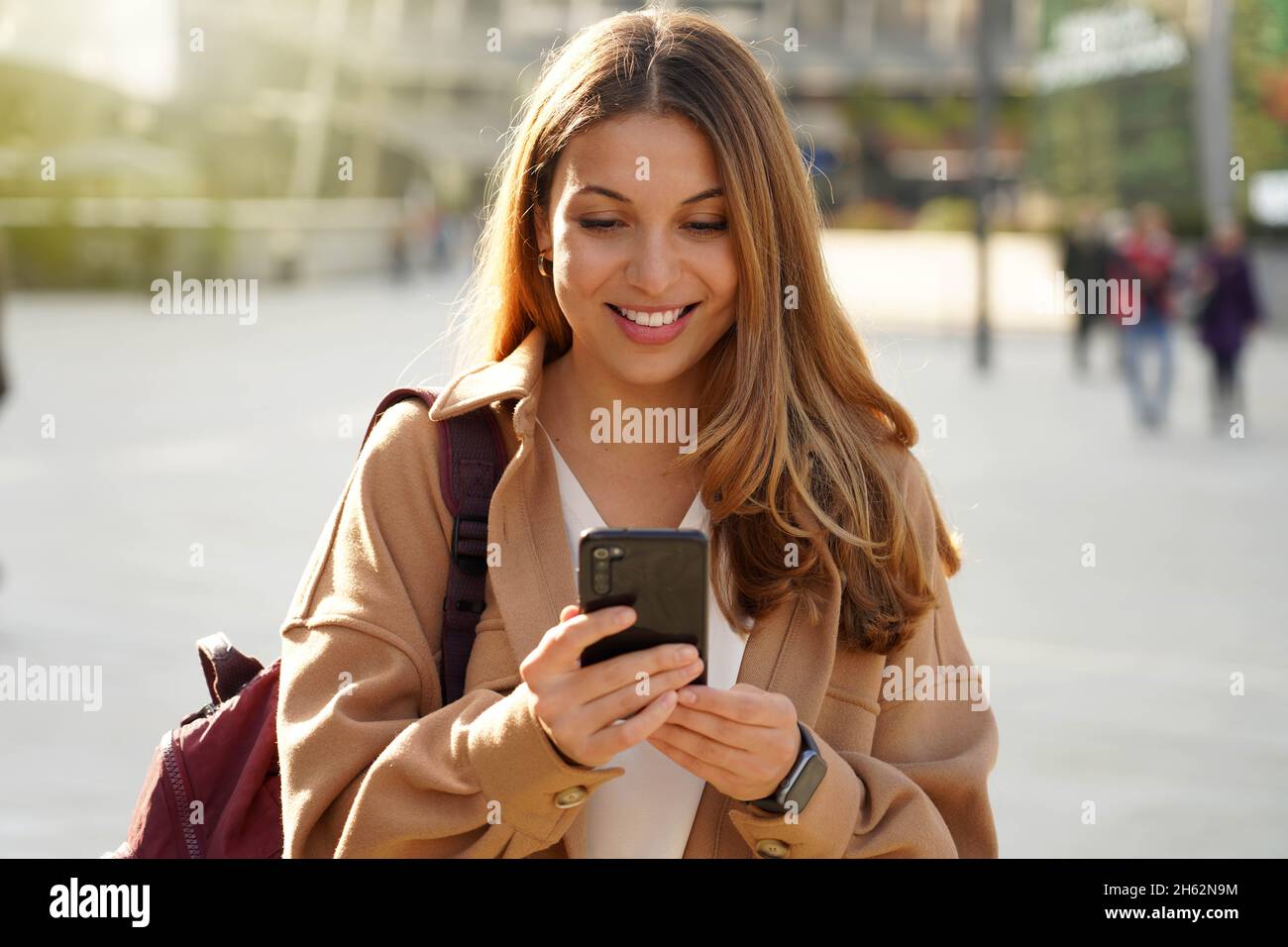 Young cheerful stylish woman using cell phone and texting message on city street. Beautiful happy casual girl holding smart phone in hands and smiling Stock Photo