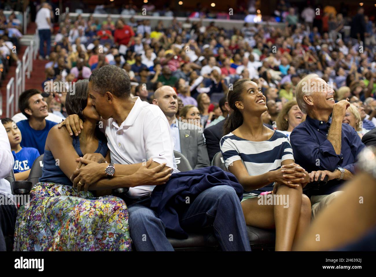 President Barack Obama kisses First Lady Michelle Obama for the 'Kiss Cam' while attending the U.S. Men's Olympic basketball team's game against Brazil at the Verizon Center in Washington, D.C., July 16, 2012. Vice President Joe Biden and Malia Obama look up at the jumbotron. Stock Photo