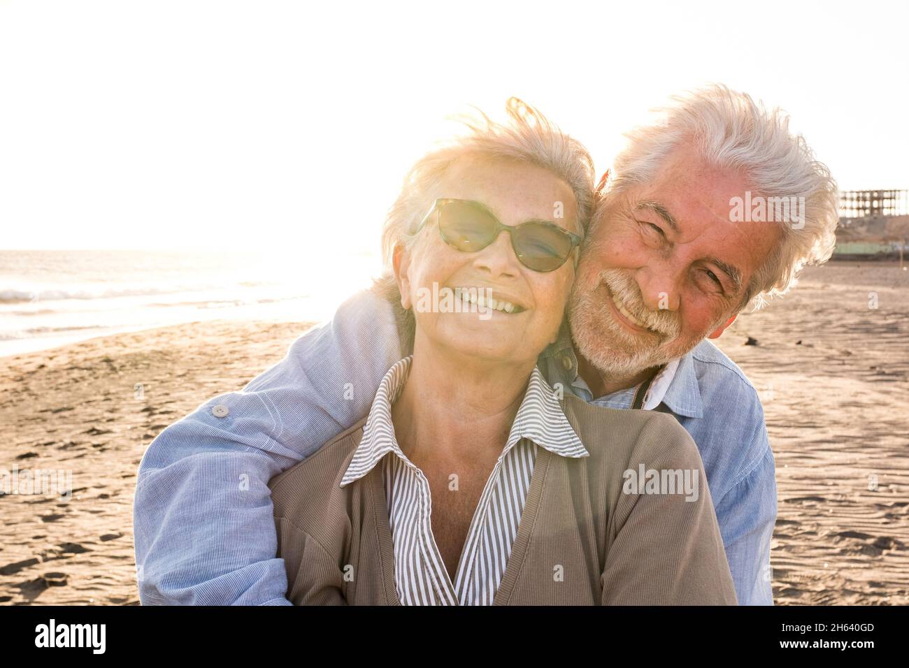 portrait of couple of mature and old people enjoying summer at the beach looking to the camera smiling and having fun together with the sunset at the background. two active seniors traveling outdoors. Stock Photo