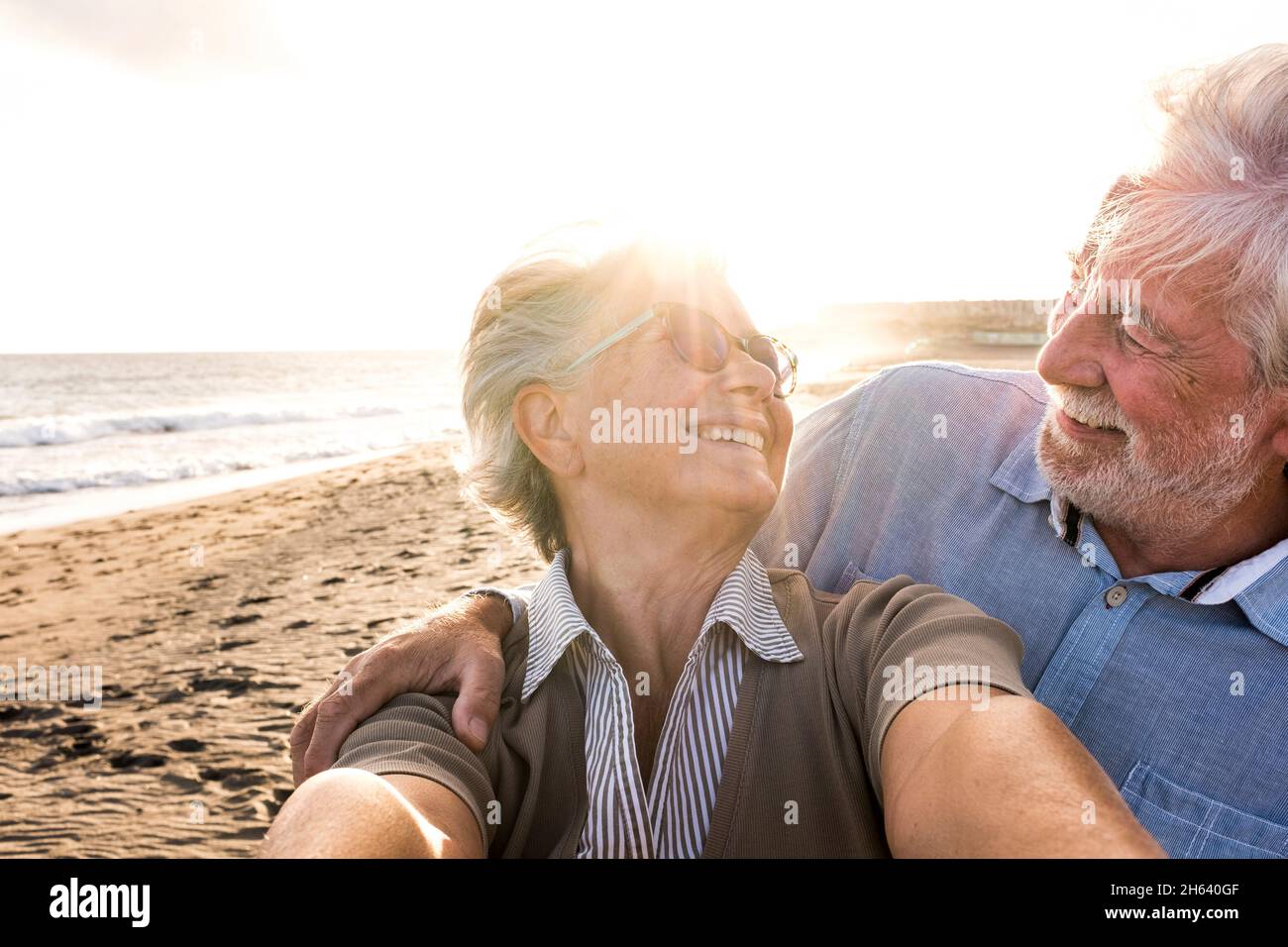 portrait of couple of mature and old people enjoying summer at the beach looking to the camera taking a selfie together with the sunset at the background. two active seniors traveling outdoors. Stock Photo