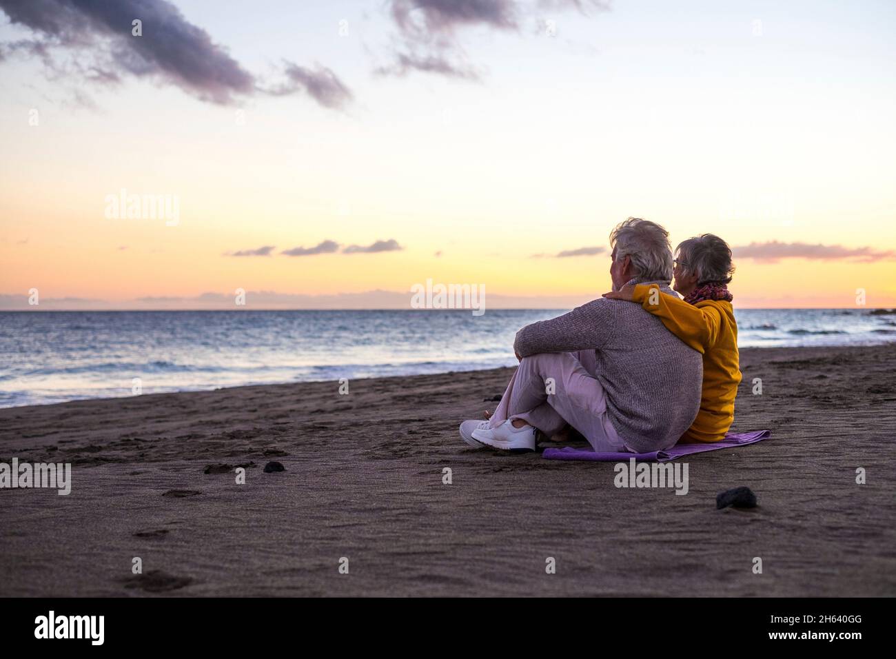 portrait of couple of mature and old people enjoying summer at the beach looking to the sea smiling and having fun together with the sunset at the background. two active seniors traveling outdoors. Stock Photo