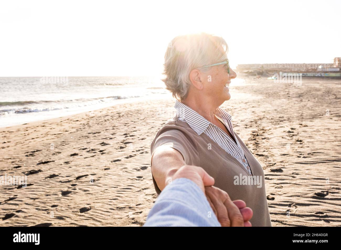 first view and pov of mature and old man holding his wife's hand at the beach having fun and enjoying together summer. two happy seniors outdoors with the sunset at the background. Stock Photo