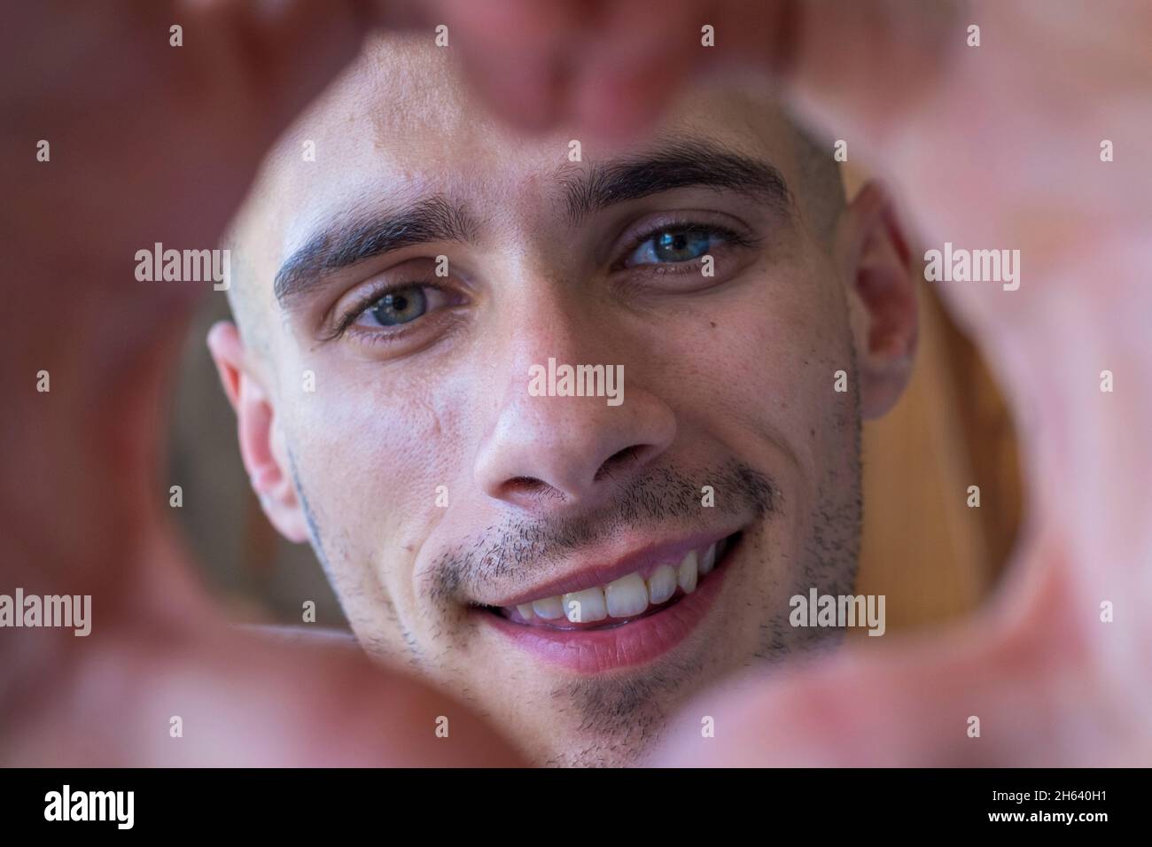 cheerful affectionate teenager man take romantic selfie portrait looking at camera through heart of joined fingers. enamored young boy showing his love to the camera doing a heart shape with his hands Stock Photo