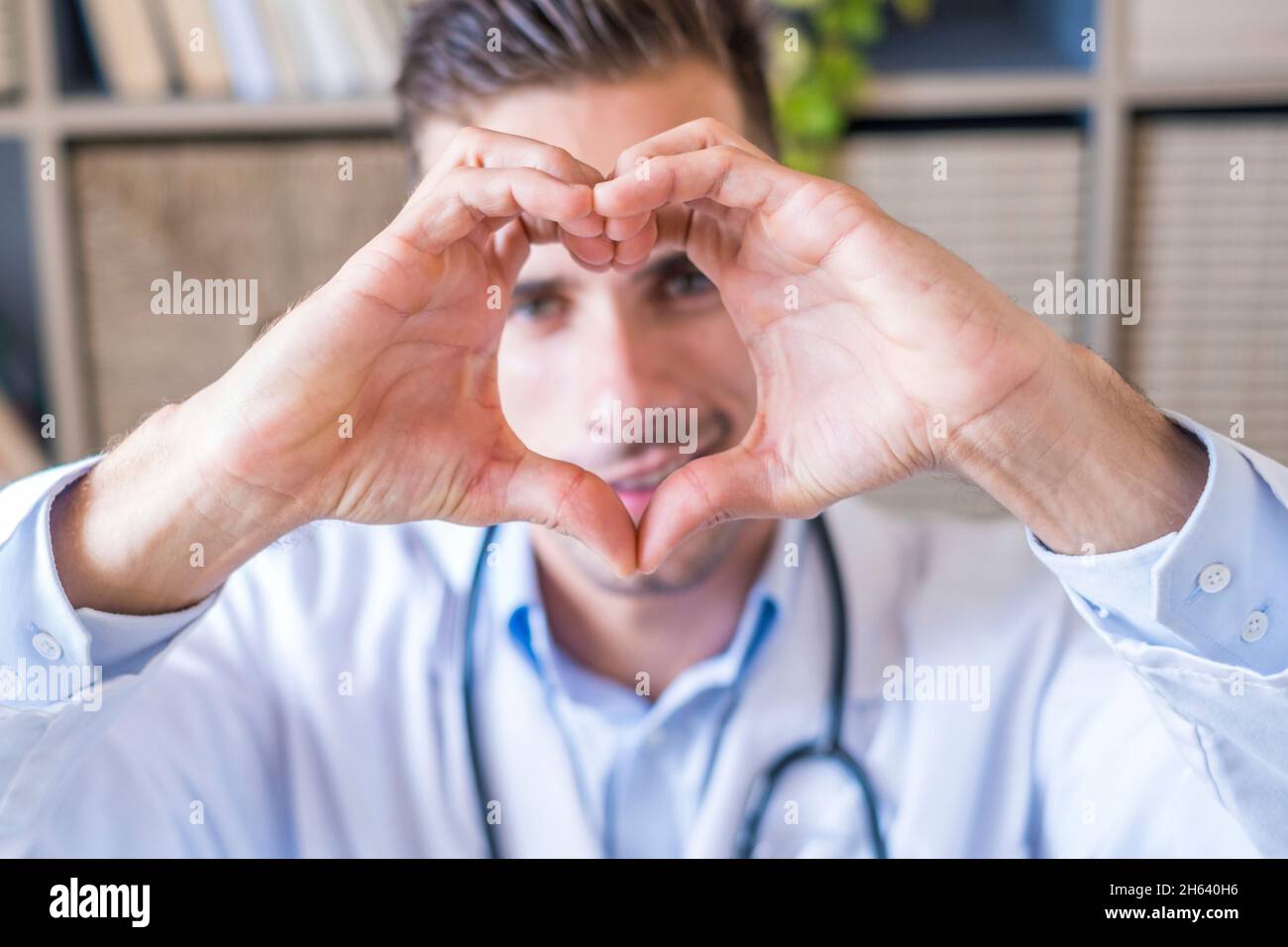 close up portrait of smiling young caucasian male nurse or gp in white medical uniform show heart love hand gesture. happy young man doctor show support and care to patients or client in hospital. Stock Photo