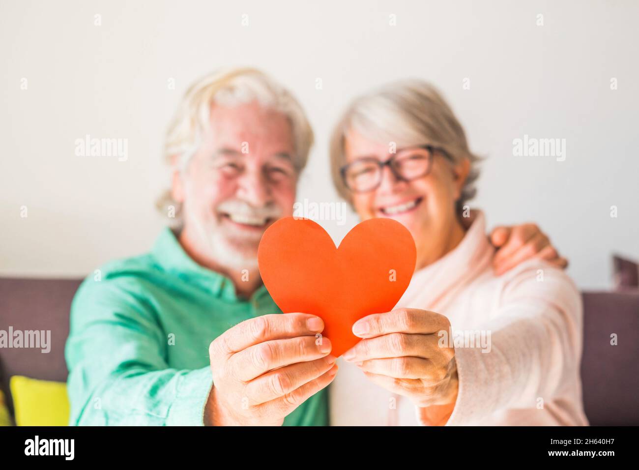 close up of two happy and in love seniors smiling and looking at the camera holding a red heart together - mature people in love Stock Photo