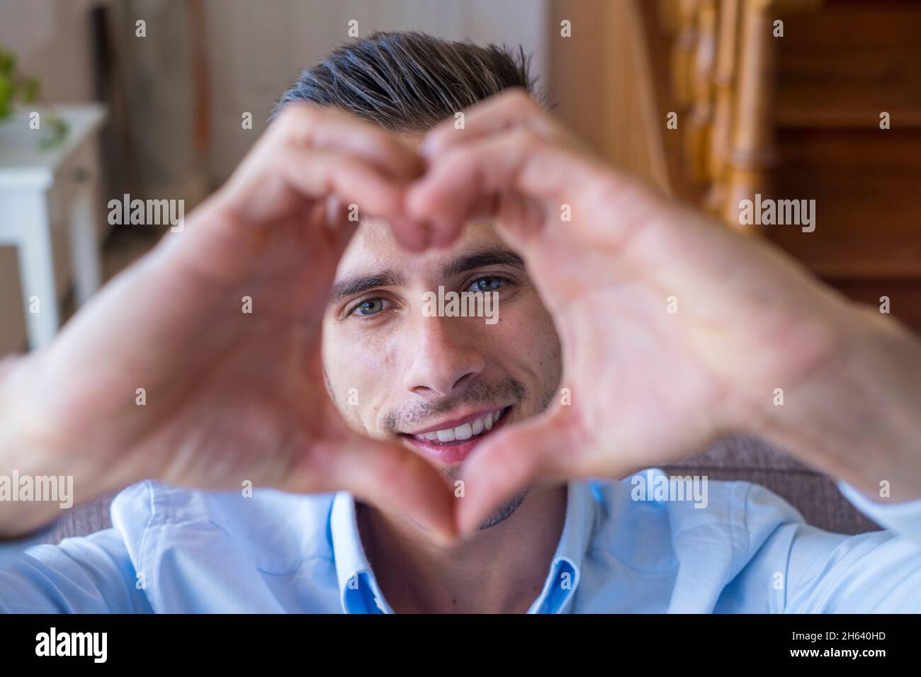 cheerful affectionate teenager man take romantic selfie portrait looking at camera through heart of joined fingers. enamored young boy showing his love to the camera doing a heart shape with his hands Stock Photo